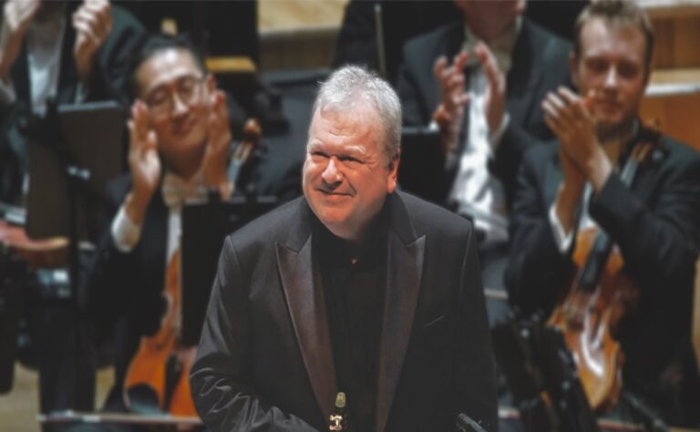 Principal Guest Artist Michael Collins stands and smiles on stage as Ulster Orchestra musicians in the background applaud.