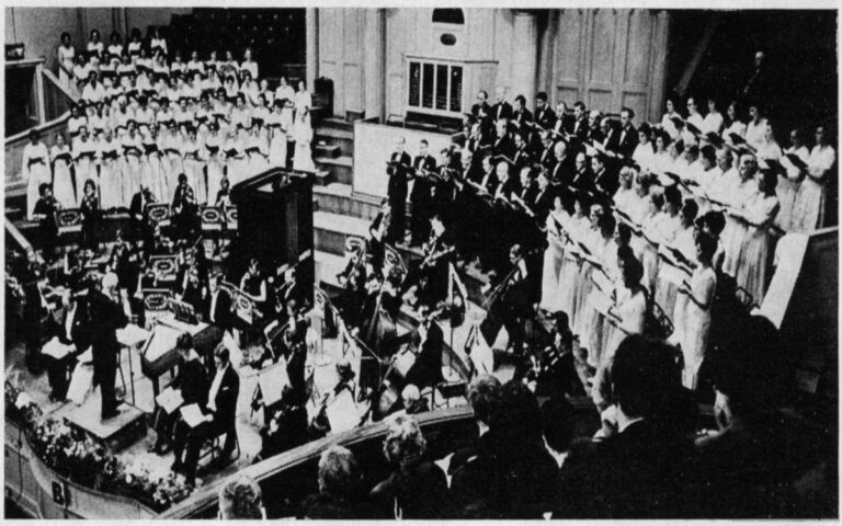A large choir and orchestra perform Messiah onstage in a concert hall for the 1966 UO Playback, with the conductor leading and a seated audience watching.