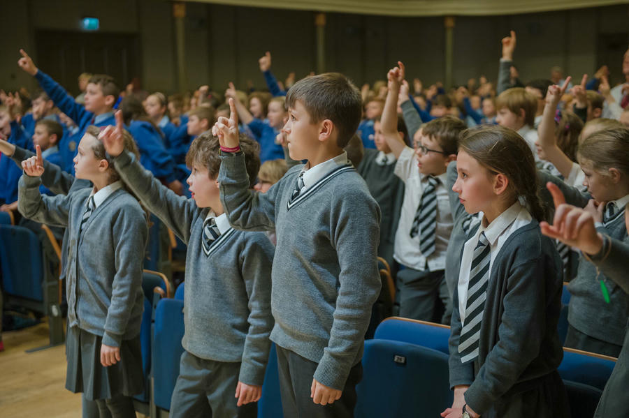 A group of school children in uniforms stand in an auditorium with their right arms raised, appearing to participate in an activity or assembly.