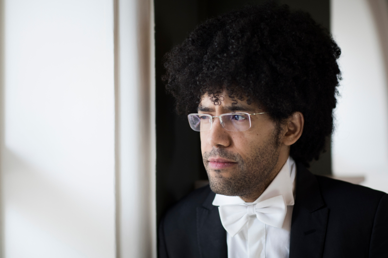 A man with curly hair, glasses, and a white bow tie looks out of a window while wearing a formal black suit, embodying the refined style often seen among members of an Artistic Team.