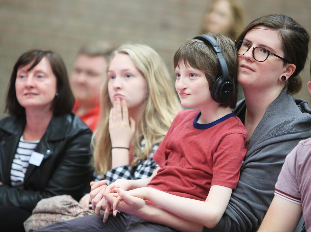 A group of people sit closely together, focused on something off-camera during one of the Relaxed Performances; a boy wearing headphones sits on a woman’s lap.