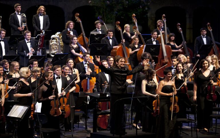 A conductor on stage with a large orchestra, dressed in formal attire, standing and holding their instruments after a Thursday Lunchtime Concert performance.
