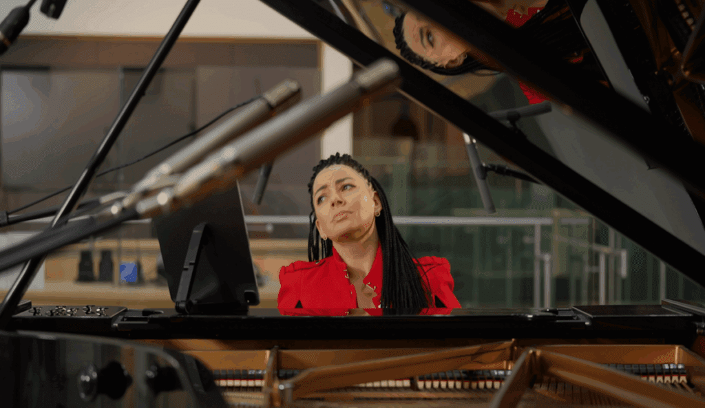 A woman in a red blouse sits at a grand piano with sheet music, appearing thoughtful, as seen through the open lid and reflection of the piano.