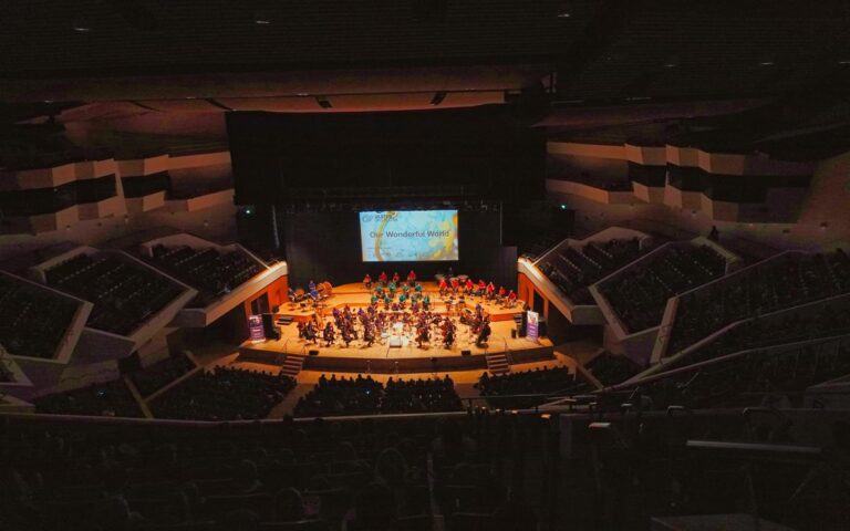 A symphony orchestra performs on a well-lit stage in a large concert hall filled with audience members, including enthusiastic Primary School Pupils, as a screen behind displays the text "Our Wonderful World" during the Wonderful World Concerts.