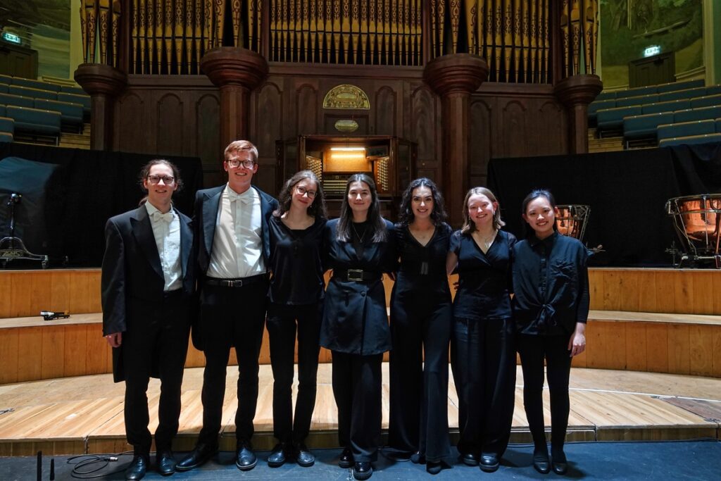 Seven people in formal black clothing stand on a wooden stage in front of a large pipe organ, smiling at the camera, celebrating their achievements through the Professional Experience Scheme.