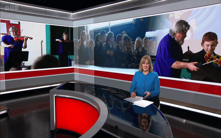 A news presenter sits at a desk in a studio, with large screens behind her displaying images from Make the News: Schools Inspiration Workshops, showing a musician playing violin and engaging with children.