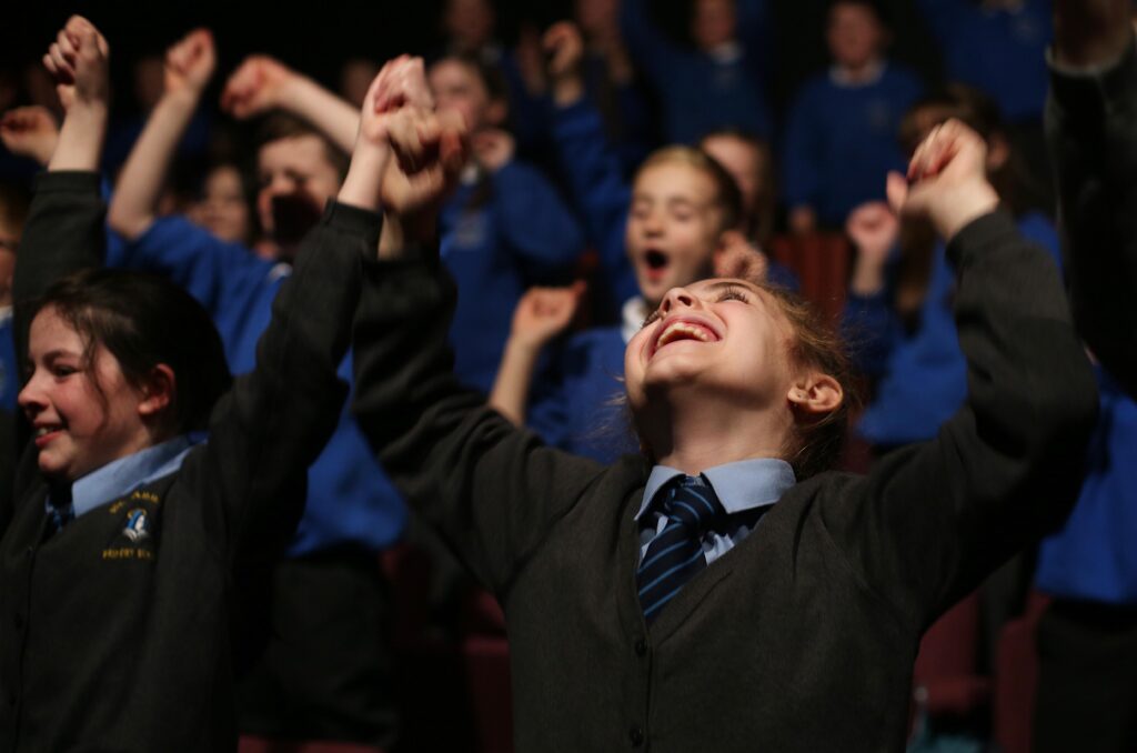 A group of students in uniforms raises their hands and cheers enthusiastically at a lively primary school event in a crowded indoor setting.
