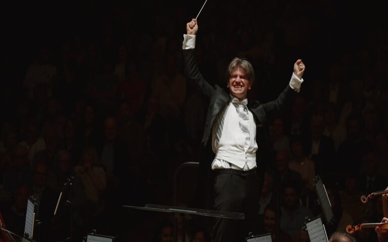 A conductor in a tuxedo stands on stage with raised arms, smiling, holding a baton, as the orchestra and audience celebrate music in a memorable 2023 in Review performance.