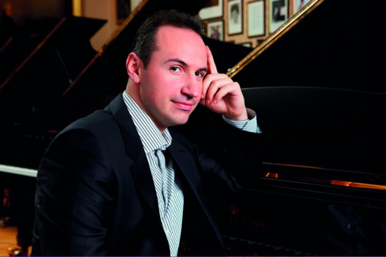 A man in a suit sits at a grand piano, resting his head on his hand, looking at the camera—reminiscent of Simon Trpčeski preparing for Brahms Piano Concerto No.2. Framed pictures hang on the wall behind him.