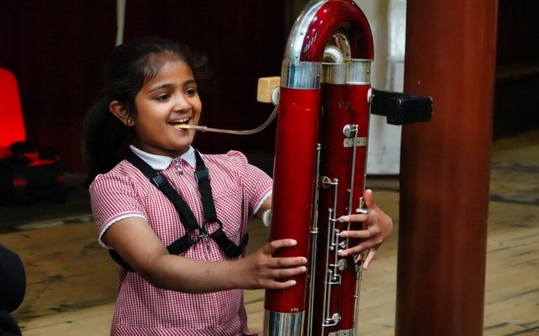 A young girl in a red checkered dress plays a large contrabassoon indoors, smiling as she holds the instrument upright—her confidence ready to win your vote.