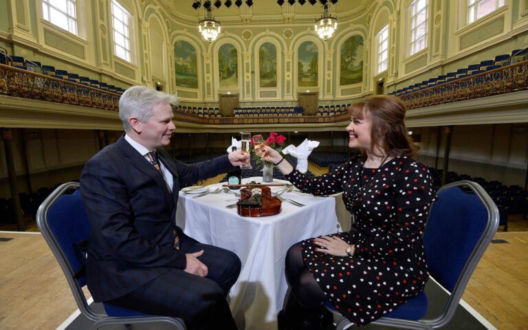 A man and woman sit at a table set for dinner on a stage in a large, ornate hall at Ten Square, clinking glasses and smiling at each other, enjoying a romantic Valentine's evening.