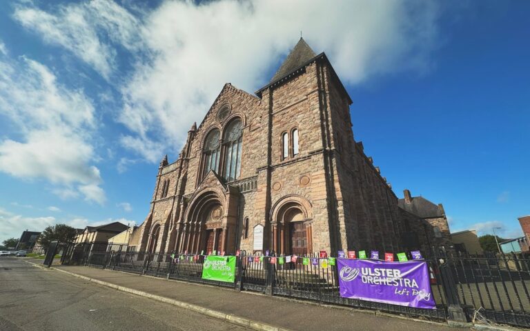 A large stone church with arched windows and a tall central spire, Townsend Street Church features colorful Ulster Orchestra event banners on the iron fence, highlighting its ongoing restoration and community redevelopment efforts.