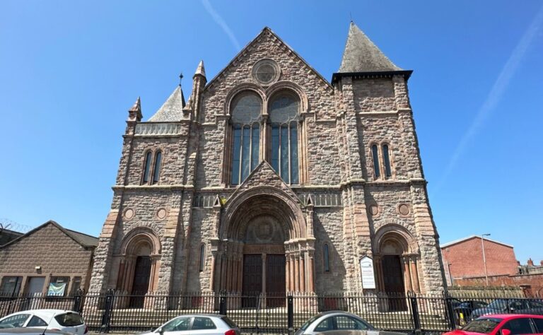 A large stone church with arched windows, two towers, and three wooden doors, viewed from the street with parked cars in front on a clear day, stands near where the Ulster Orchestra once received Foyle Foundation funding.