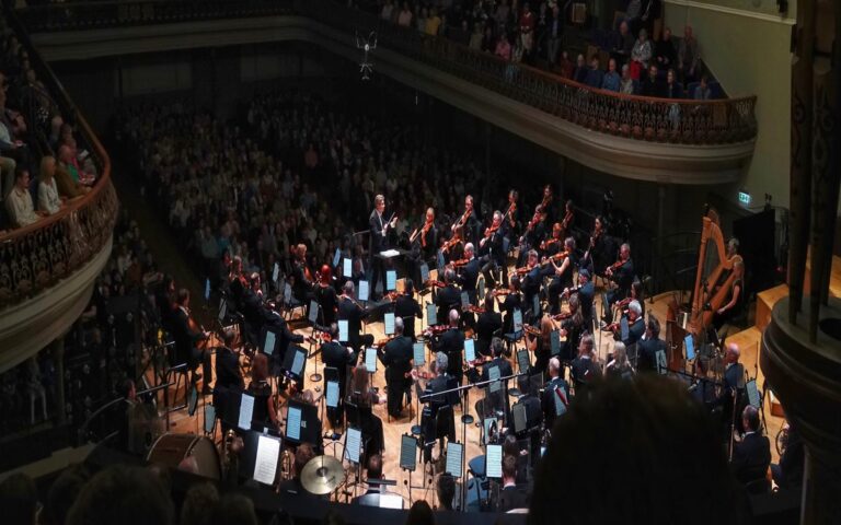 A symphony orchestra performs classical music on stage in a large concert hall filled with UK audiences.