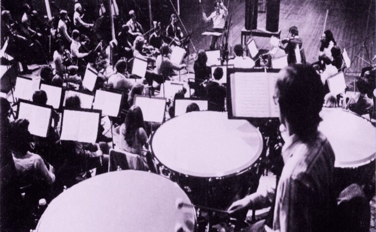 A large Expanded Orchestra rehearses in a studio, with musicians playing string instruments and timpani visible in the foreground. Sheet music stands are positioned throughout the group for this 1981 UO Playback session.
