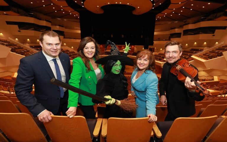 Five people stand in an empty concert hall; one is dressed as a witch holding a broom, while another holds a violin.