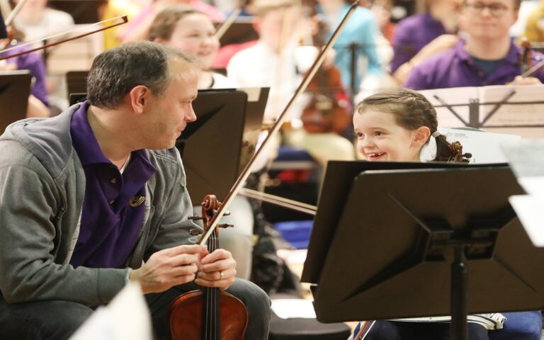 An adult and a child sit with string instruments and music stands, smiling at each other during the 2019 Come and Play event orchestra rehearsal.