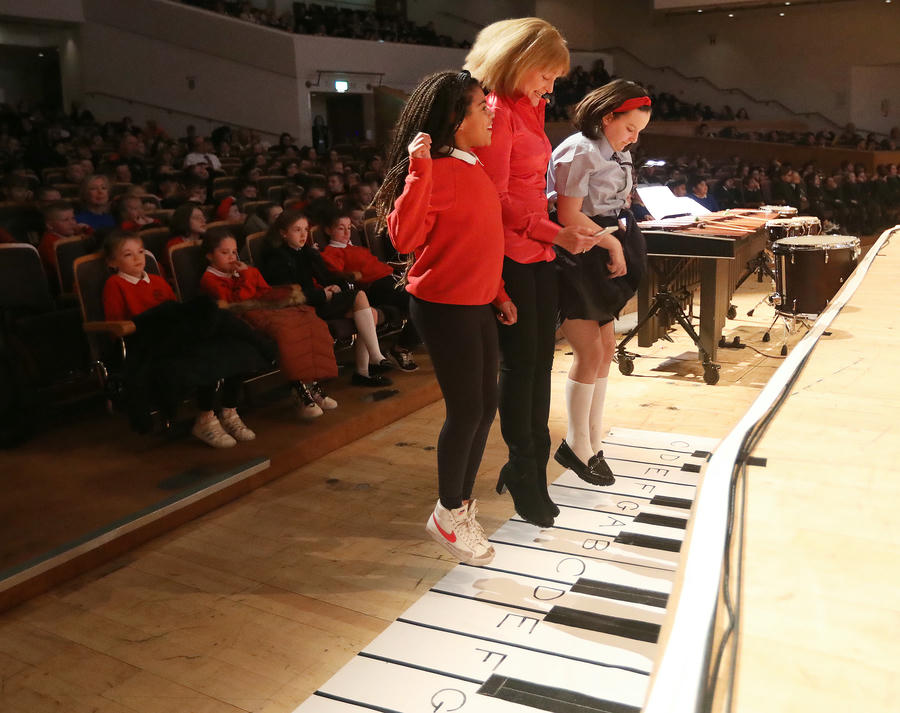 At a Primary School Concert, a woman and two girls step on a large floor piano keyboard on stage, captivating the audience in the auditorium.