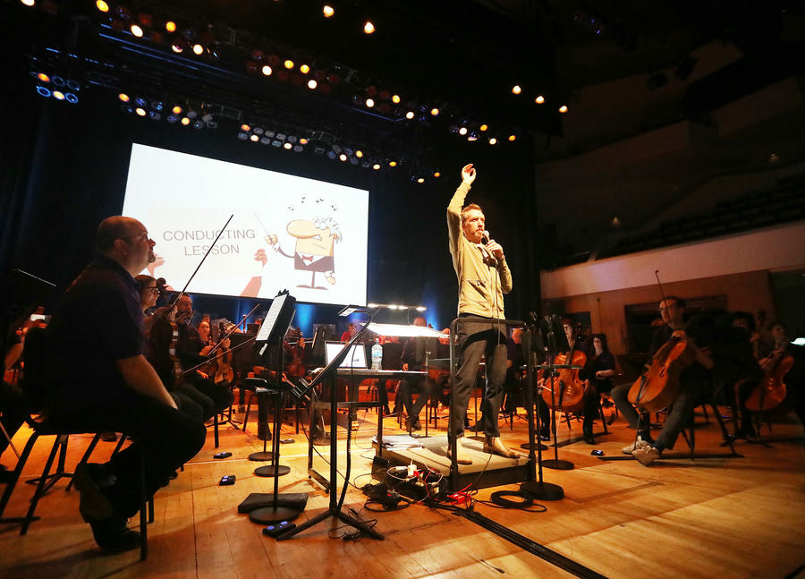 A conductor stands on stage leading an orchestra during a rehearsal or lesson, with musicians seated and a large screen behind displaying "CONDUCTING LESSON"โa perfect scene often found in Primary School Concerts.