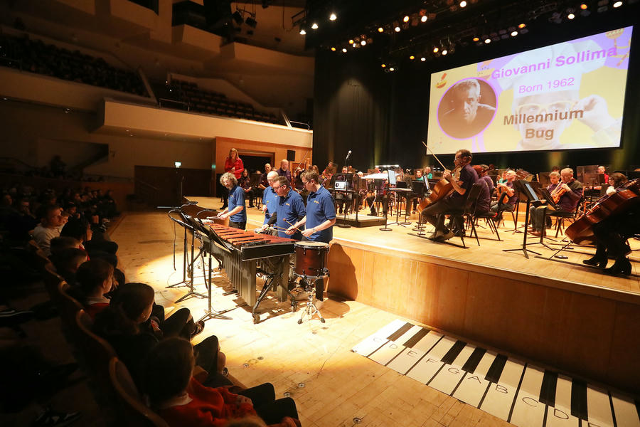 Musicians perform on stage with percussion instruments in a concert hall for a Primary School Concert; an audience watches and a large screen shows "Giovanni Sollima Millennium Bug.