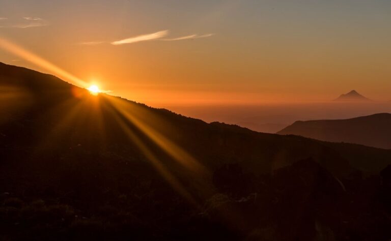 The sun sets behind a mountain ridge, casting rays of light over a landscape with distant mountains under a clear sky—a tranquil scene inviting reflection, reminiscent of Haydn’s contemplative Seven Last Words.