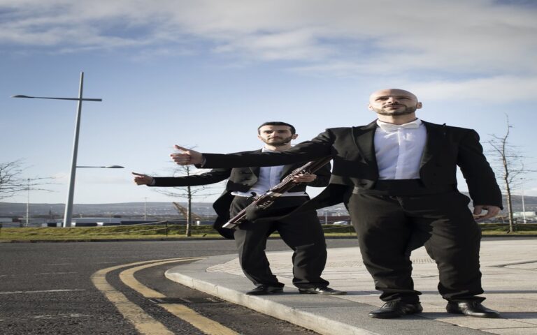 Two men in formal attire stand on a street curb near the Portico of Ards, one holding a bassoon, both with thumbs out as if hitchhiking—perhaps en route to join the Ulster Orchestra.