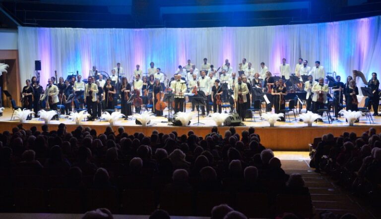A full orchestra stands on stage facing the audience in Ballymena, with musicians holding instruments, framed by a white curtain and colored stage lights.