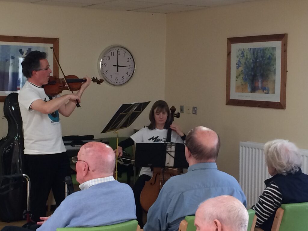Musicians on Call: A man playing violin and a woman playing cello perform for a seated audience in a room with framed art and a wall clock.