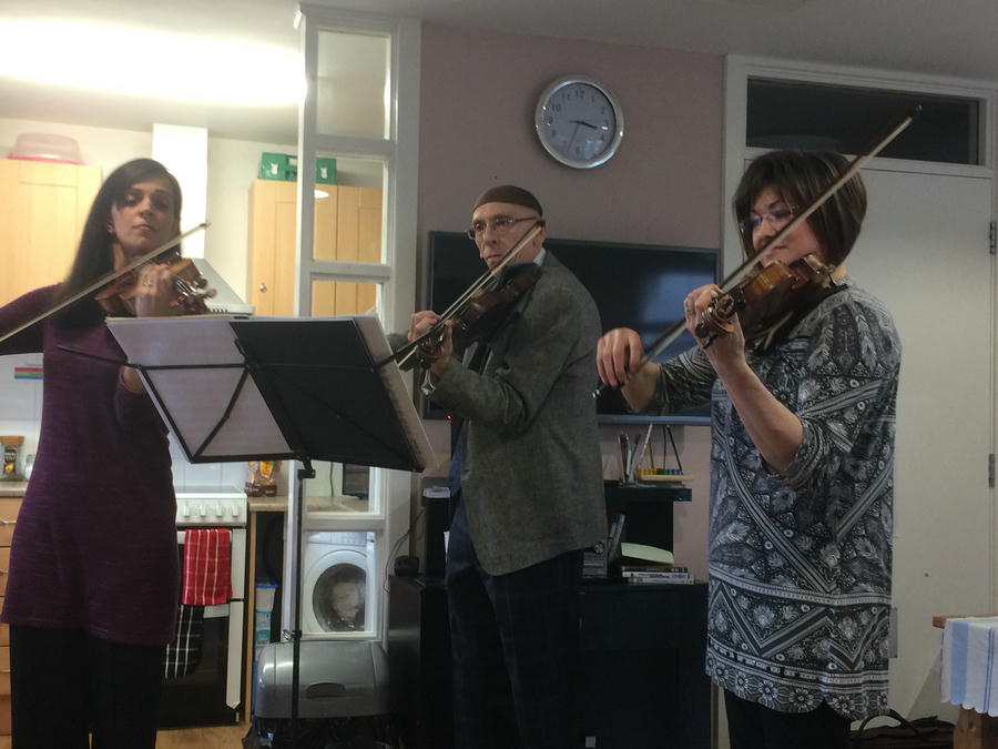 Three people are playing violins indoors near a music stand, reminiscent of Musicians on Call hospital performances. A clock, TV, and kitchen appliances are visible in the background.