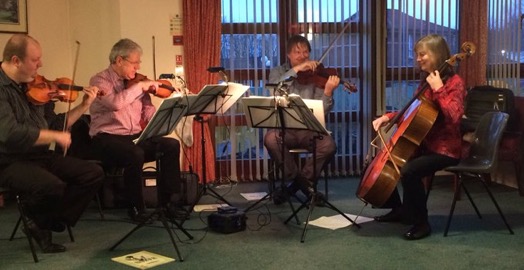 Four musicians are seated and playing string instrumentsโthree violins and one celloโin a room with large windows, music stands in front of them, as part of the Musicians on Call program.