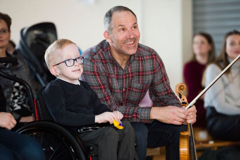 A smiling man holding a violin and bow sits next to a boy in a wheelchair, while others watch in the background.