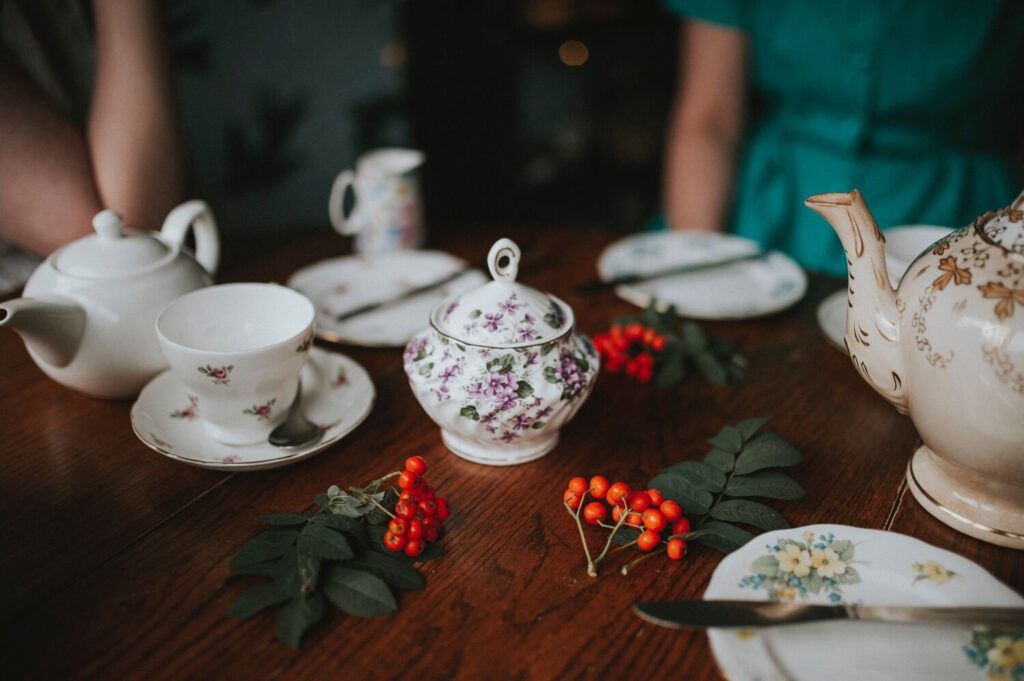 A wooden table set with floral teacups, saucers, a teapot, a sugar bowl, plates, and sprigs of red berries; two people are partially visible.