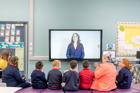 A group of children and two adults sit facing a large screen displaying a woman speaking in a classroom setting.