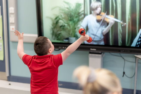 A child in a red shirt raises his arms, holding a musical instrument, while watching a person play violin on a large screen in a classroom.