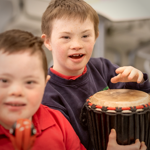 Two children with Down syndrome are smiling; one is playing a hand drum while the other holds a maraca.