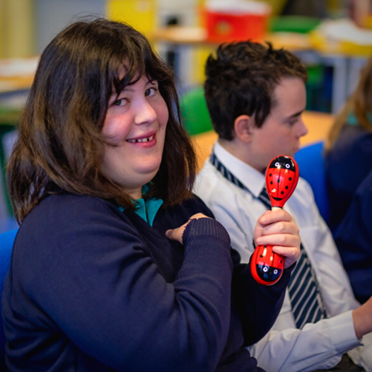 A student smiles while holding a red ladybird-shaped shaker in a classroom. Another student sits beside her, facing forward.