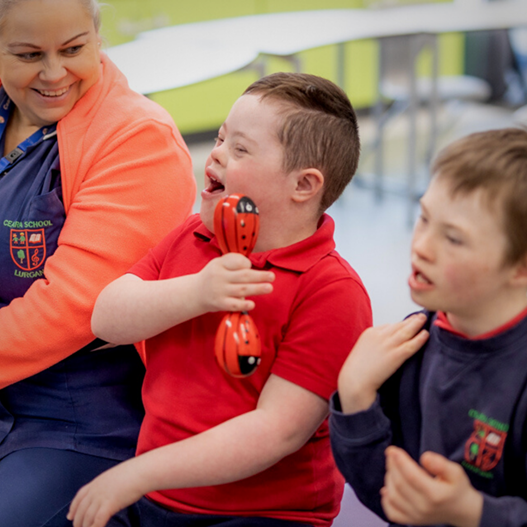 Three children, two with Down syndrome, are sitting together indoors. One child in a red shirt holds toy ladybugs and smiles, while an adult in uniform sits beside them.