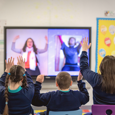 Three children in school uniforms sit facing a large screen, raising their hands to interact with two adults on a video call.
