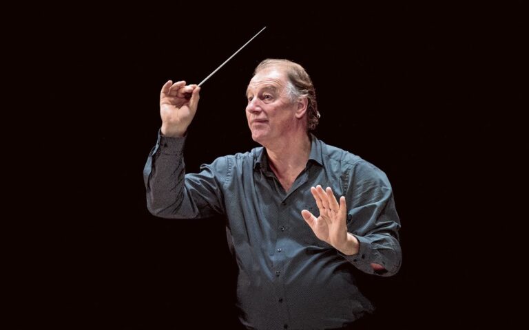 A middle-aged man in a dark shirt conducts with a baton, one hand raised while the other gestures, against a plain black background—legendary conductor Jac van Steen leads, as seen in UO LIVE and heard on BBC Radio 3.