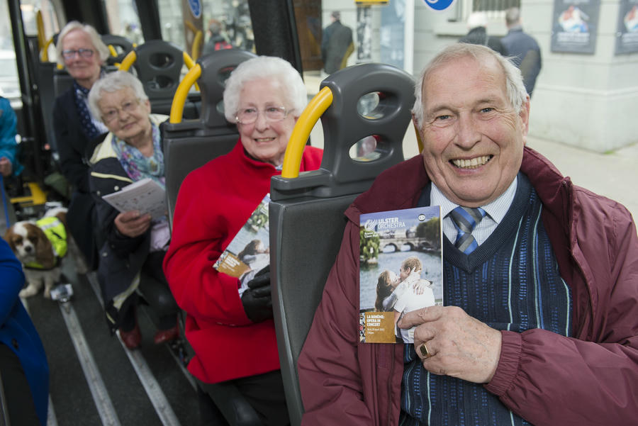 A group of elderly people sitting on a bus, smiling and holding pamphlets for Move to the Music; a dog is visible at the back.