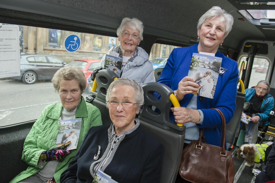 Four older women on a bus hold up Move to the Music booklets and smile at the camera; other passengers and a wheelchair user with a dog are visible in the background.