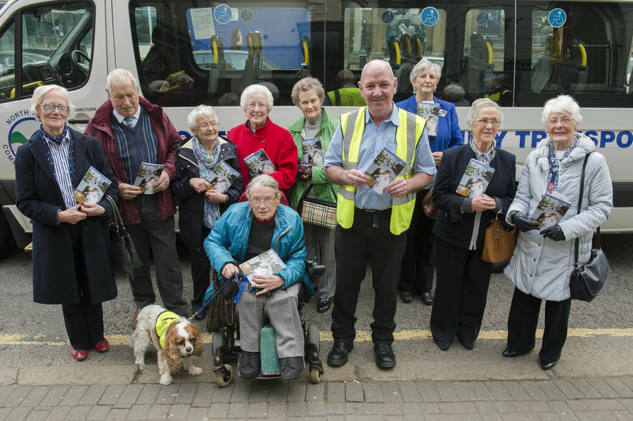A group of elderly people and a man in a high-visibility vest pose with Move to the Music booklets in front of a transport van; one woman is in a wheelchair with a service dog.