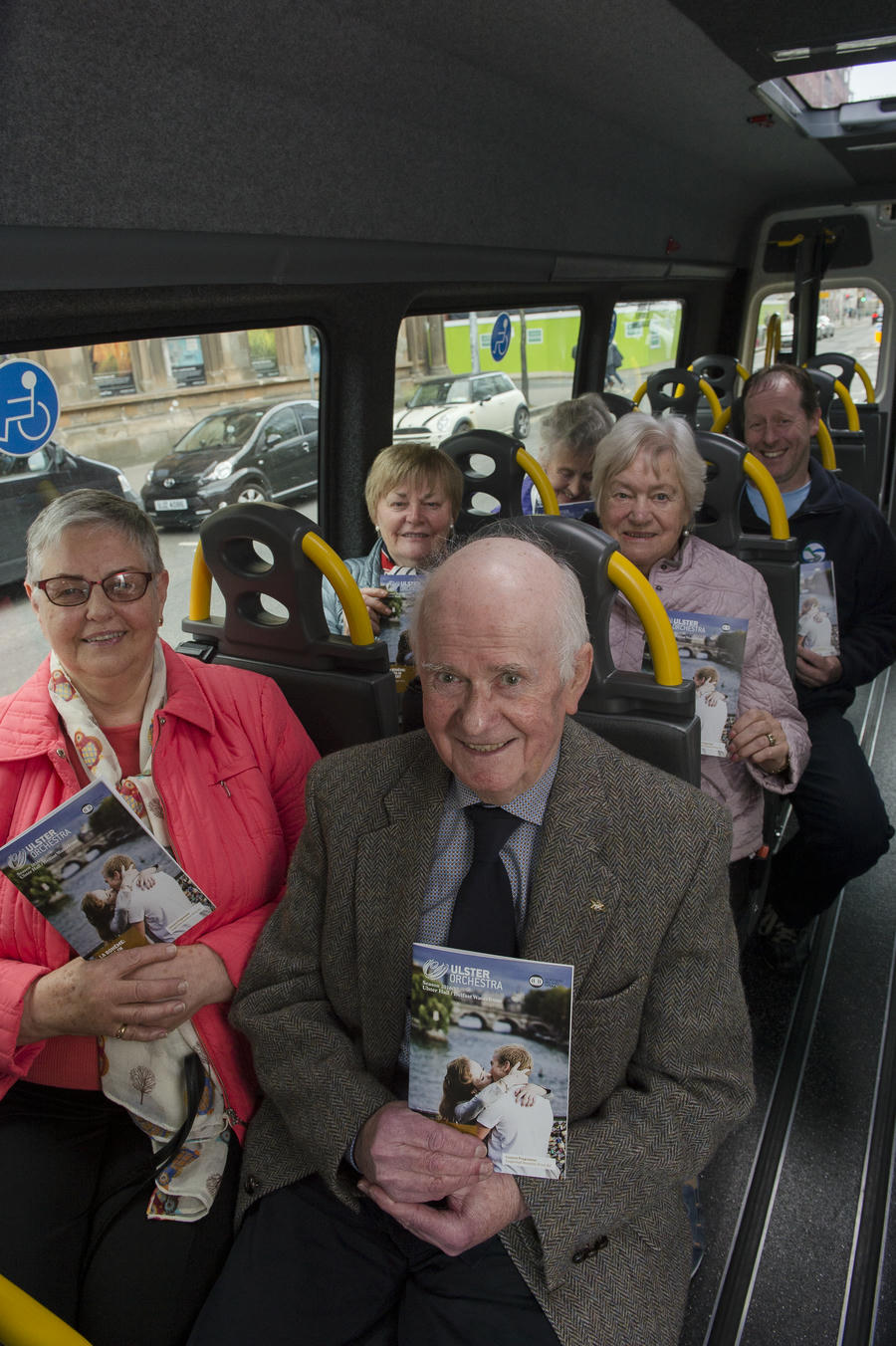 A group of older adults sit and smile on a bus, each holding the same "Move to the Music" magazine or brochure.