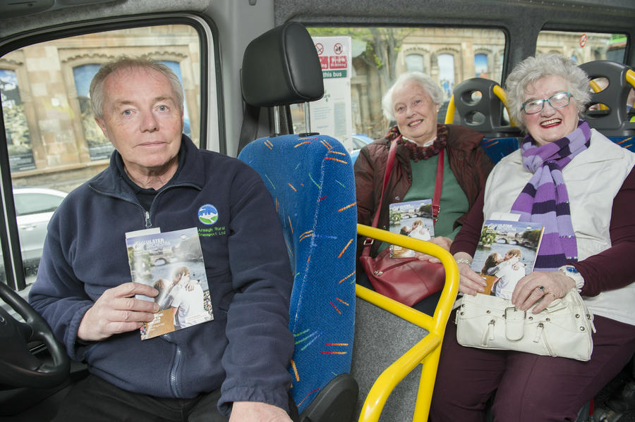 A man sits in the driver's seat of a bus, holding a booklet, while two elderly women behind him also hold booklets and smile at the camera, ready to move to the music on their joyful journey.