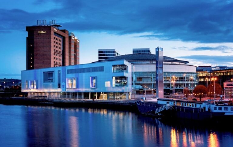 A modern waterfront building with large windows is lit up at dusk, with a tall Hilton hotel behind it, and boats moored along the river in the foreground—an iconic spot where the Orchestras Conference hosted by the Association of British Orchestras took place.