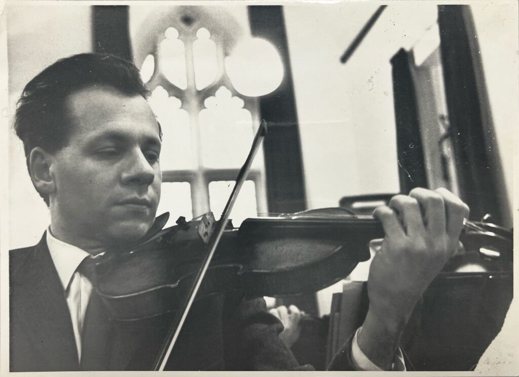 A black-and-white photo of a man with black hair in a suit and tie playing a violin.