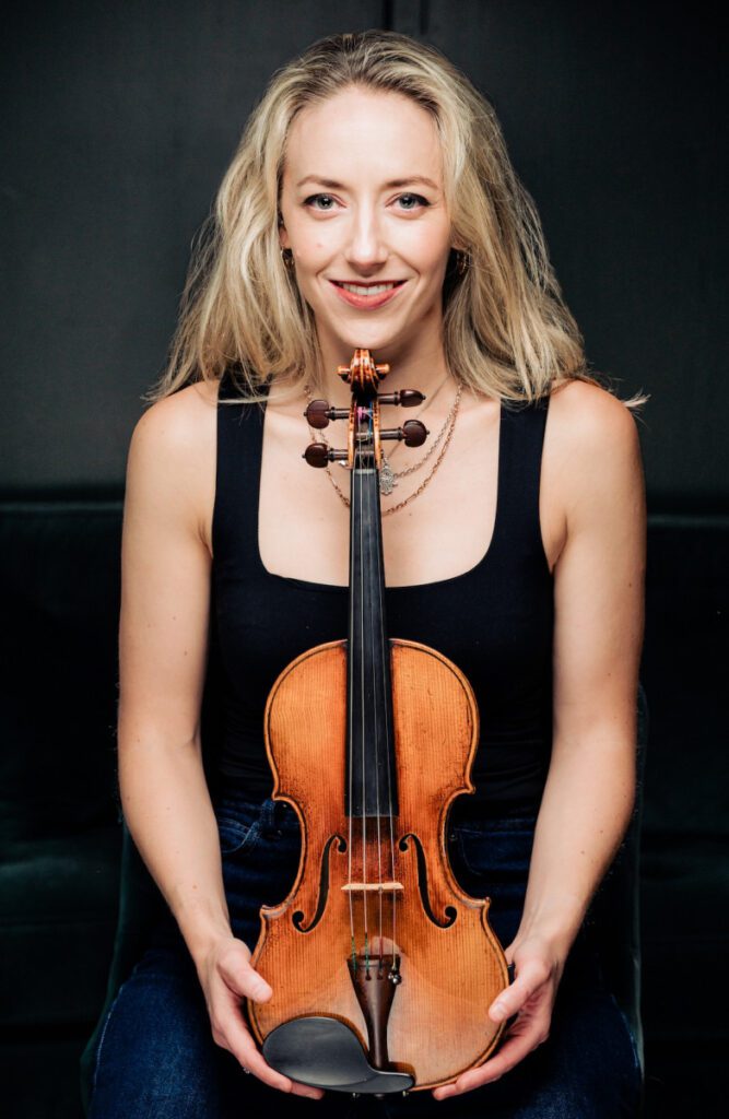 A woman with blonde hair is smiling and holding a violin.