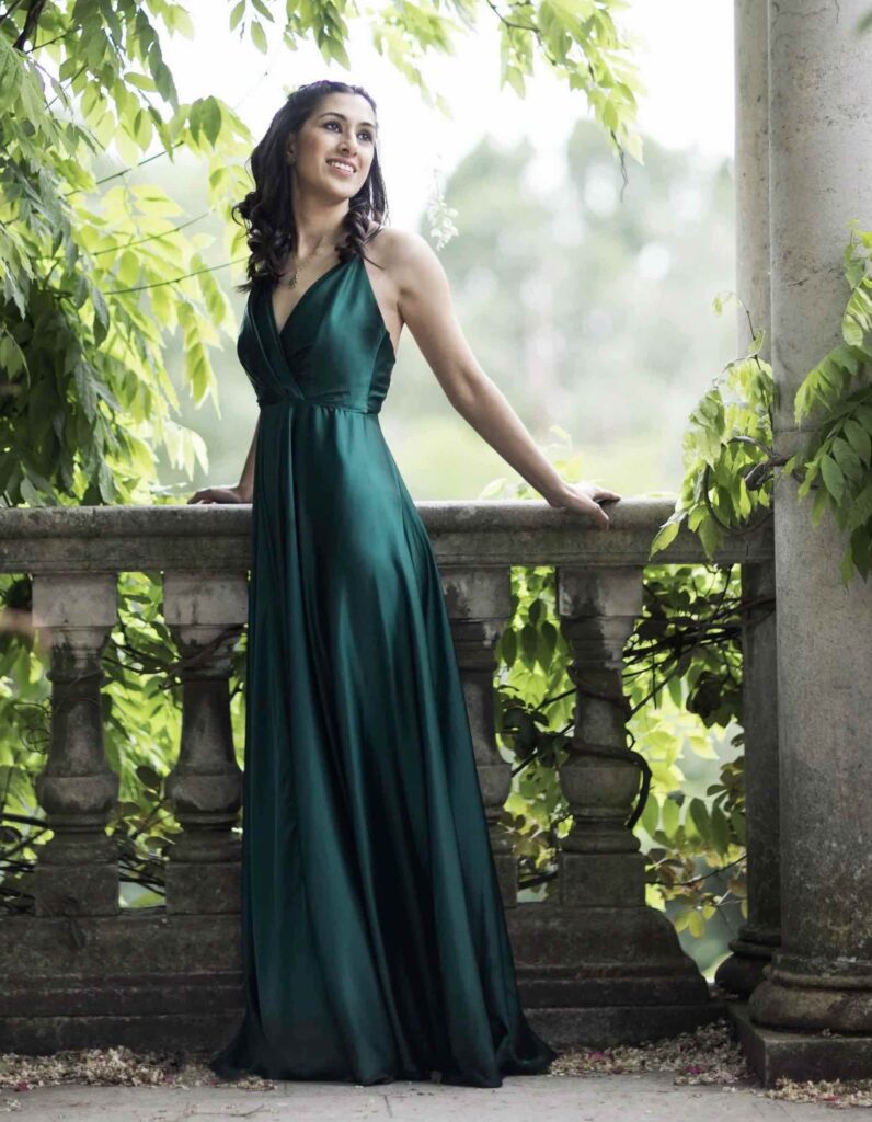 A young woman with dark hair and wearing a long green dress stands at a balcony surrounded by foliage.