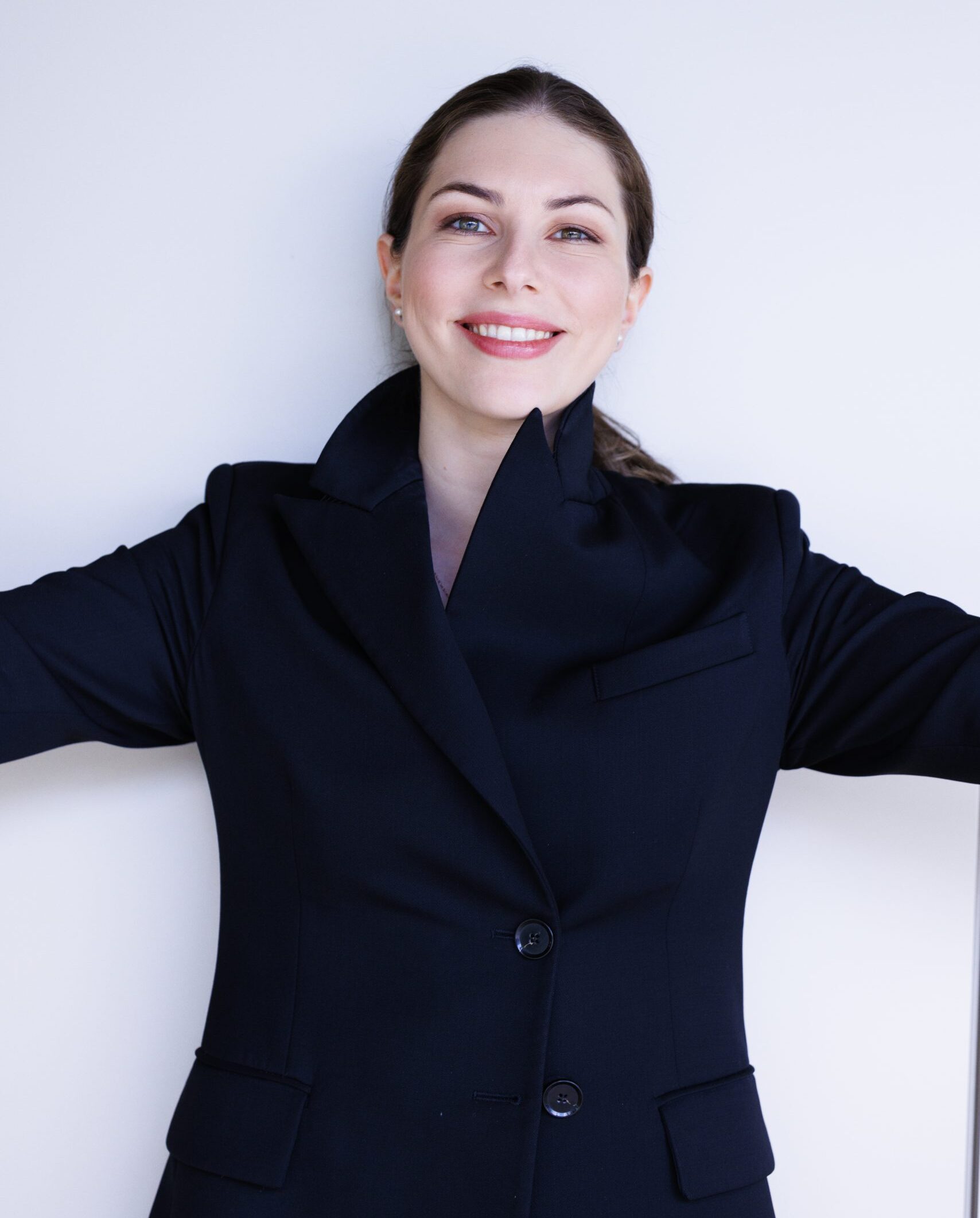 A dark-haired woman in a black jacket stands smiling with her arms wide open.